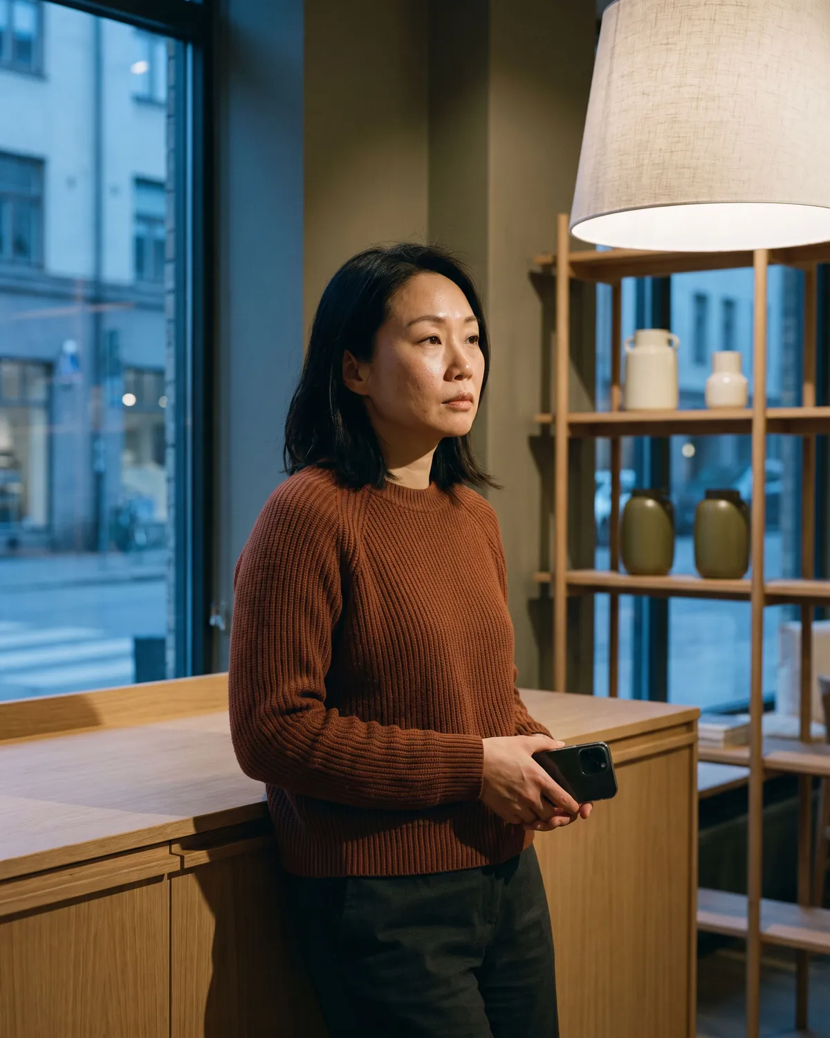 A small-business owner stands in her studio after closing, phone in hand, still waiting for a call back.